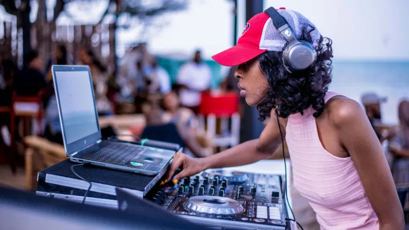 Person wearing headphones and a red cap is DJing at an outdoor event with a laptop and mixer, while a crowd gathers in the background, showcasing perfect gift ideas for musicians.