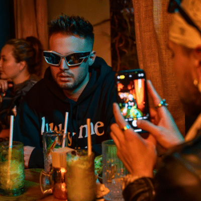 A man wearing sunglasses sits at a table with drinks, while another person snaps his photo with a smartphone—perhaps capturing content on how to make money on TikTok in the dimly lit setting.