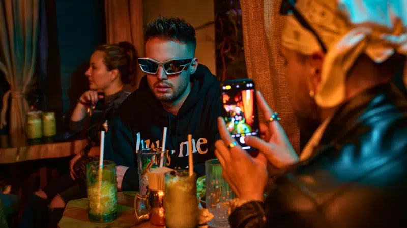 A man wearing sunglasses sits at a table with drinks, while another person snaps his photo with a smartphone—perhaps capturing content on how to make money on TikTok in the dimly lit setting.