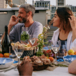 Submit Your Music To Our Dinner Table Jazz Playlist On Spotify Five people sit around a rooftop table set with food, drinks, and flowers, enjoying lively conversation and a daytime meal as a Jazz Spotify playlist sets the perfect mood. Buildings are visible in the background.