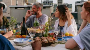 Five people sit around a rooftop table set with food, drinks, and flowers, enjoying lively conversation and a daytime meal as a Jazz Spotify playlist sets the perfect mood. Buildings are visible in the background.