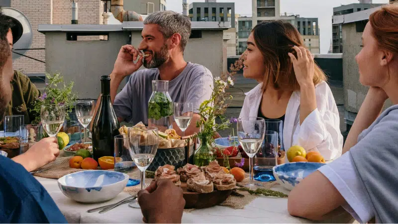 Submit Your Music To Our Dinner Table Jazz Playlist On Spotify Five people sit around a rooftop table set with food, drinks, and flowers, enjoying lively conversation and a daytime meal as a Jazz Spotify playlist sets the perfect mood. Buildings are visible in the background.