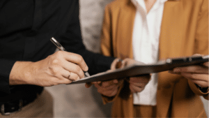 6 Types Of Record Label Contracts Two people stand side by side; one holds a clipboard while the other signs contracts with a pen. Faces are not visible.