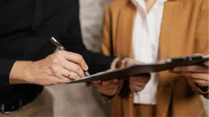 Two people stand side by side; one holds a clipboard while the other signs contracts with a pen. Faces are not visible.