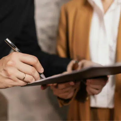 Two people stand side by side; one holds a clipboard while the other signs contracts with a pen. Faces are not visible.
