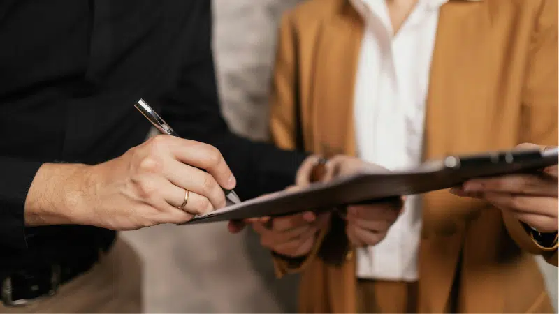 Two people stand side by side; one holds a clipboard while the other signs contracts with a pen. Faces are not visible.