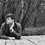 A man sitting on a dock in the woods, enjoying the peaceful ambiance while listening to fresh new music.