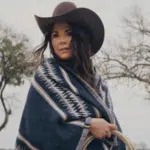 A woman donning a stylish cowboy hat and a cozy blanket poses gracefully amidst a vast field, exuding an air of serenity.