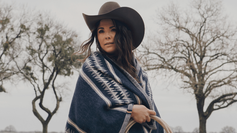 A woman donning a stylish cowboy hat and a cozy blanket poses gracefully amidst a vast field, exuding an air of serenity.