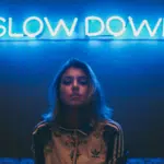 A person with shoulder-length hair stands in front of a blue neon sign that says "SLOW DOWN," capturing the essence of a music industry slowdown.