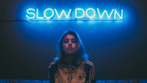 A person with shoulder-length hair stands in front of a blue neon sign that says "SLOW DOWN," capturing the essence of a music industry slowdown.