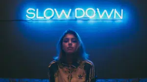 A person with shoulder-length hair stands in front of a blue neon sign that says "SLOW DOWN," capturing the essence of a music industry slowdown.