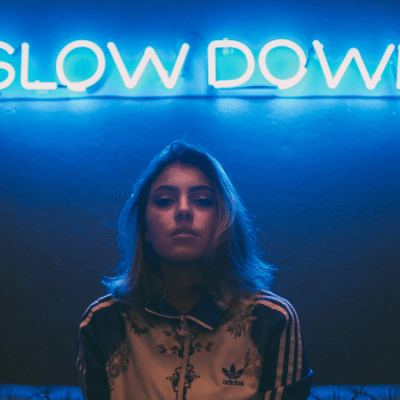 A person with shoulder-length hair stands in front of a blue neon sign that says "SLOW DOWN," capturing the essence of a music industry slowdown.