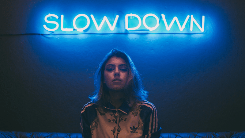 A person with shoulder-length hair stands in front of a blue neon sign that says "SLOW DOWN," capturing the essence of a music industry slowdown.