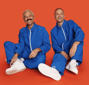 Two men in blue jumpsuits sitting on a red background.