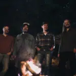 A fresh group of men standing in front of a campfire, enjoying new music.