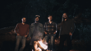 A fresh group of men standing in front of a campfire, enjoying new music.