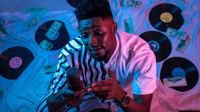 A man in a striped shirt holds a microphone, surrounded by vinyl records and scattered dollar bills, as if he's just received grants for musicians.