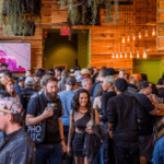 A large crowd gathers inside a wooden-paneled venue for a music conference, with people chatting and holding drinks. Banners with "Symphonic" are displayed in the background.