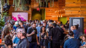 A large crowd gathers inside a wooden-paneled venue for a music conference, with people chatting and holding drinks. Banners with "Symphonic" are displayed in the background.