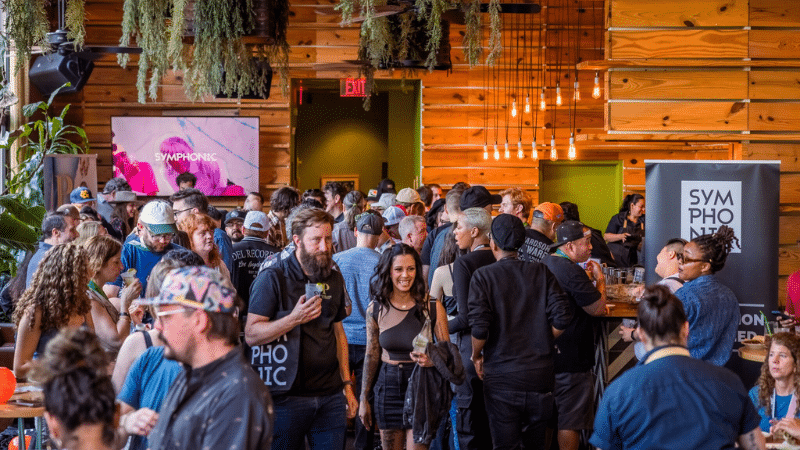 A large crowd gathers inside a wooden-paneled venue for a music conference, with people chatting and holding drinks. Banners with "Symphonic" are displayed in the background.