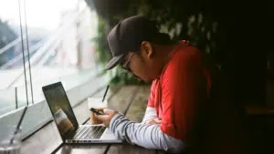 A man in a red shirt and cap sits at a wooden table with a laptop and a drink, looking at his smartphone near a large window, likely reading music industry newsletters.