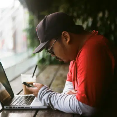 A man in a red shirt and cap sits at a wooden table with a laptop and a drink, looking at his smartphone near a large window, likely reading music industry newsletters.