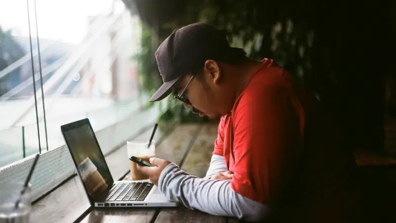 A man in a red shirt and cap sits at a wooden table with a laptop and a drink, looking at his smartphone near a large window, likely reading music industry newsletters.