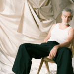 A man sitting on a chair with fresh new music playing in front of a curtain.
