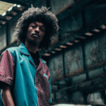 A person with curly hair stands under a rusted metal bridge, wearing a patterned shirt with blue and red colors, looking at the camera while listening to a Spotify playlist.