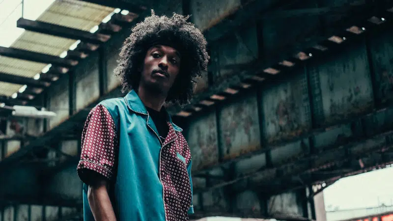 A person with curly hair stands under a rusted metal bridge, wearing a patterned shirt with blue and red colors, looking at the camera while listening to a Spotify playlist.