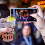 Person taking a photo of a crowded venue with a smartphone, while holding a beverage in the foreground.