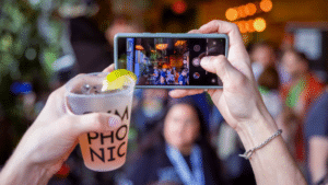 Person taking a photo of a crowded venue with a smartphone, while holding a beverage in the foreground.