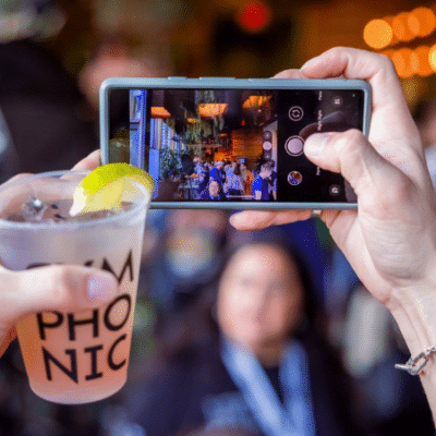 Person taking a photo of a crowded venue with a smartphone, while holding a beverage in the foreground.