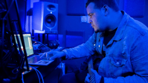 Man working on music production in a studio with a guitar and laptop.