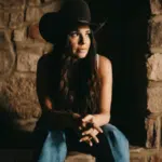 Woman in a cowboy hat sitting against a stone wall, headphones on, listening to fresh new music.