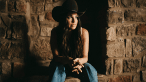 Woman in a cowboy hat sitting against a stone wall, headphones on, listening to fresh new music.