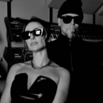 Stylish duo wearing sunglasses posing in a room filled with synthesizers, ready to create fresh new music.