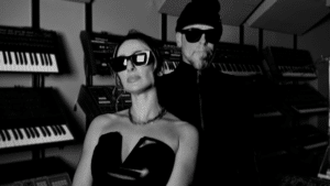 Stylish duo wearing sunglasses posing in a room filled with synthesizers, ready to create fresh new music.