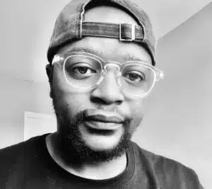 Black and white close-up of a man wearing glasses and a baseball cap facing the camera.