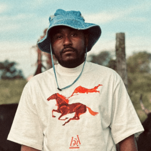 A man wearing a blue bucket hat and a white t-shirt with a red horse graphic stands in front of a blurred natural background.