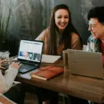 Three women sitting at a table with laptops, engaged in a pleasant conversation about empowerment.