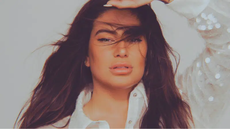 A woman with tousled, unfiltered hair wearing a white blouse, partially lit by warm light.