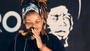 Women-Led Music Organizations You Need To Know About A woman with dreadlocks and a headwrap passionately sings into a microphone, eyes closed, in front of a black backdrop featuring a large face graphic. Celebrating women-led music, her soulful performance captures the essence of female artistry on stage.