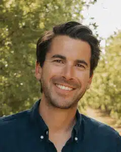 A smiling man with dark hair outdoors with trees in the background.