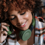 Smiling young woman with curly hair and headphones around her neck playlisting on a green phone in a bookstore.