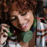 Smiling young woman with curly hair and headphones around her neck playlisting on a green phone in a bookstore.