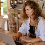 A woman with curly hair focusing on her laptop at a wooden table in a bright, rustic room, planning strategies for recoupment.