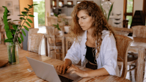 A woman with curly hair focusing on her laptop at a wooden table in a bright, rustic room, planning strategies for recoupment.