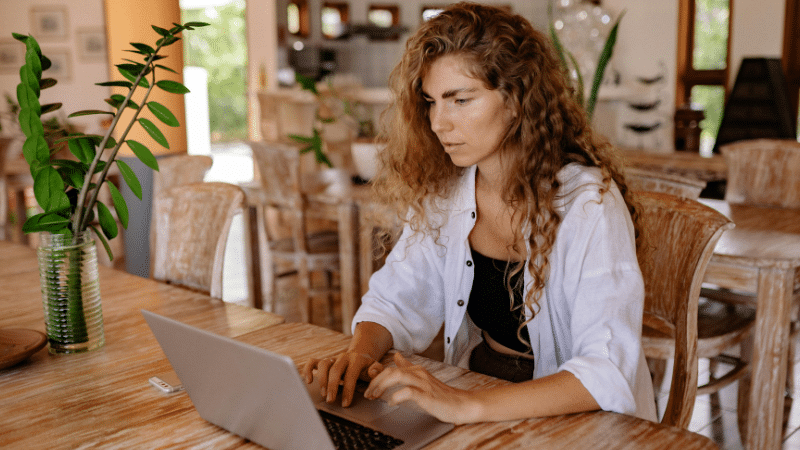 A woman with curly hair focusing on her laptop at a wooden table in a bright, rustic room, planning strategies for recoupment.